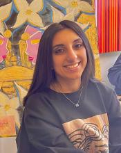 Photo of young woman, sitting with a painting in the background. 