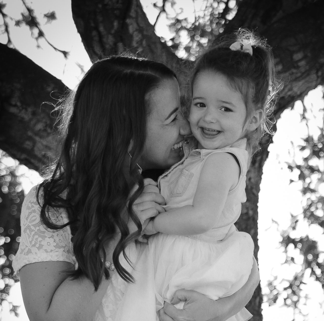 Photo in greyscale of woman with toddler. The background is a tree with light coming through. Both the woman and her child are grinning.