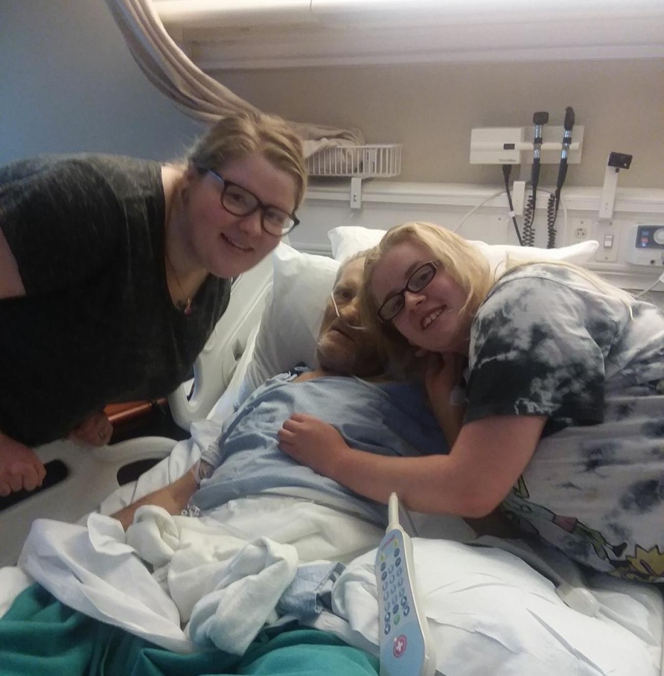 Lilly and her sister are comforting their dad in the hospital, where he lies in the bed